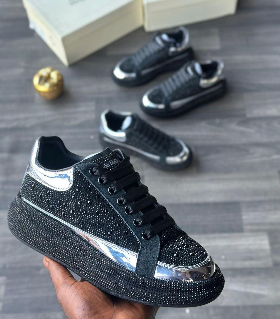 A close-up of a hand holding a black rhinestone sneaker with silver accents, with two more pairs in the background on a grey wooden floor.