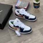 A pair of white, navy, and gold Nike Air Jordan 1 sneakers on a wooden floor, with a shoe box and game in the background.