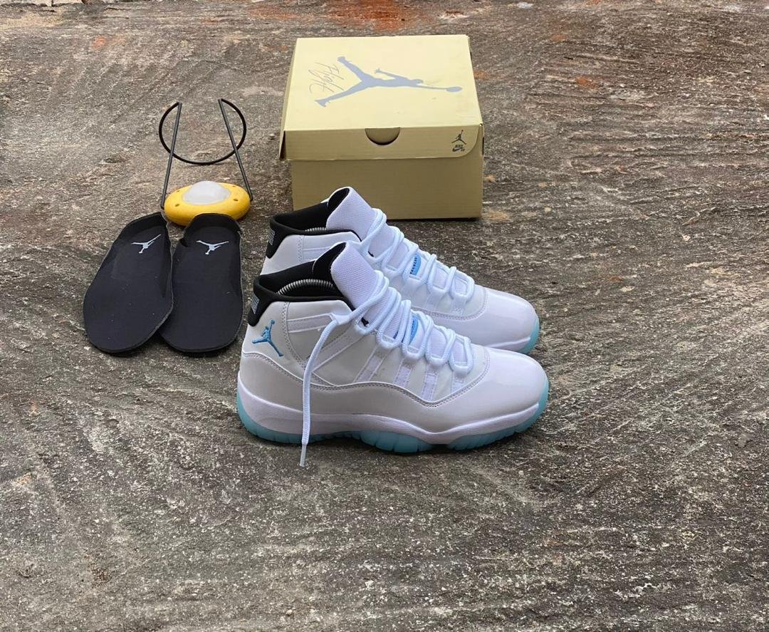 A pair of white and blue Air Jordan 11 sneakers with a yellow shoe box and black slippers on a concrete floor, perfect for sneaker enthusiasts in Nigeria.