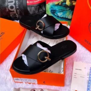 A pair of black leather slide sandals with a gold buckle, displayed in an orange box with accessories.