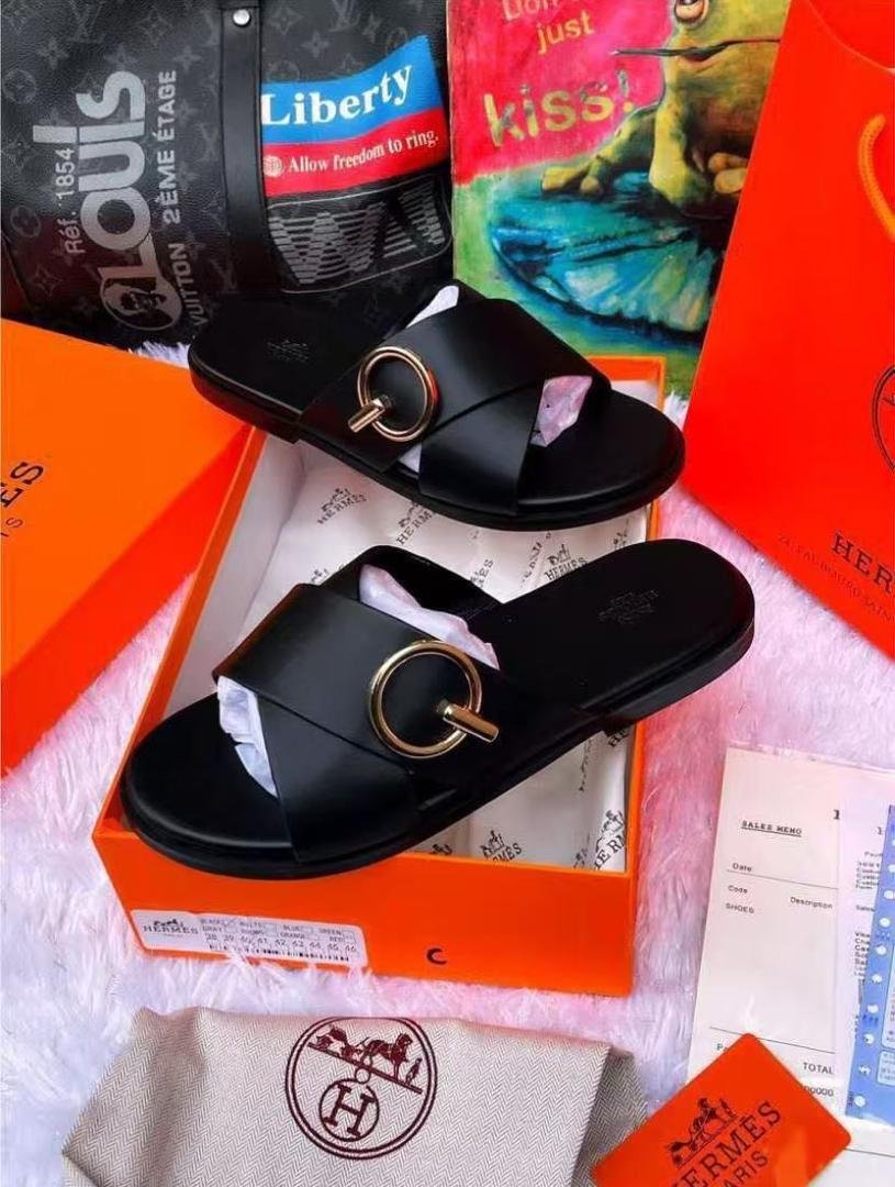 A pair of black leather slide sandals with a gold buckle, displayed in an orange box with accessories.