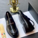 A pair of black patent leather loafers with a gold buckle on a white paper, accompanied by a shoe horn and a decorative ship's wheel.