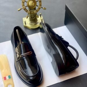 A pair of black patent leather loafers with a gold buckle on a white paper, accompanied by a shoe horn and a decorative ship's wheel.