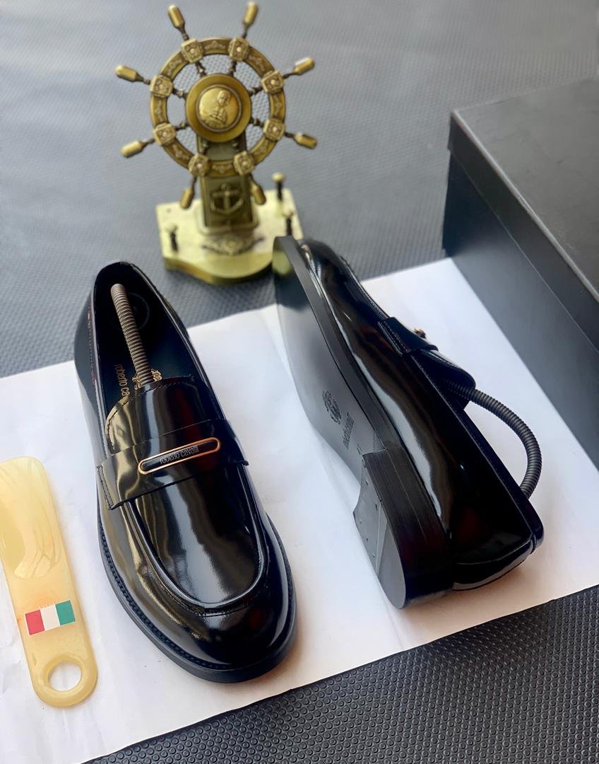 A pair of black patent leather loafers with a gold buckle on a white paper, accompanied by a shoe horn and a decorative ship's wheel.