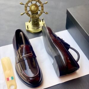 A pair of black patent leather loafers with a gold buckle on a white paper, accompanied by a shoe horn and a decorative ship's wheel.