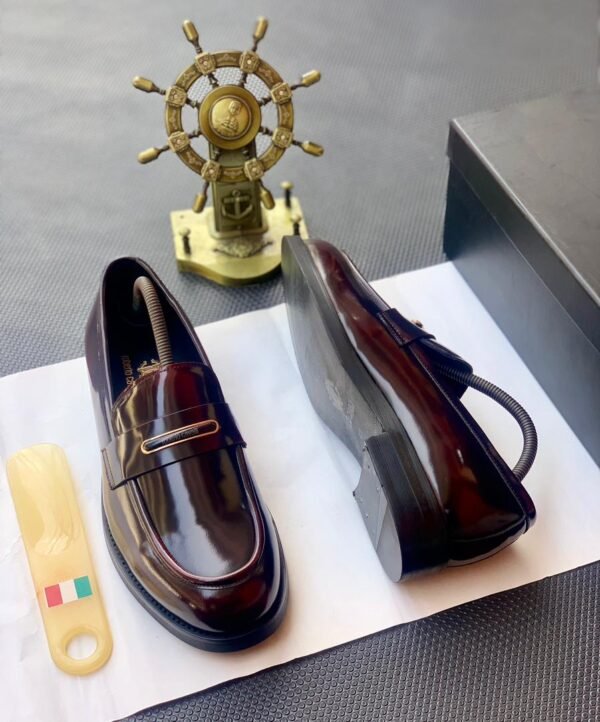 A pair of black patent leather loafers with a gold buckle on a white paper, accompanied by a shoe horn and a decorative ship's wheel.