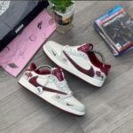 A pair of white and red Nike Air Jordan Stussy x Fragment Design sneakers on a wooden floor, surrounded by gaming and lifestyle accessories.