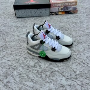 A pair of white and grey Air Jordan 4 sneakers with black accents, displayed on a marble surface with a shoe box and designer book covers in the background.
