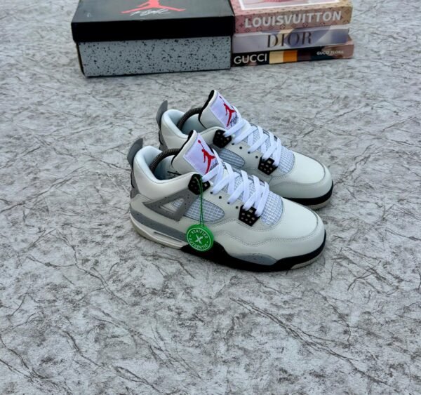 A pair of white and grey Air Jordan 4 sneakers with black accents, displayed on a marble surface with a shoe box and designer book covers in the background.