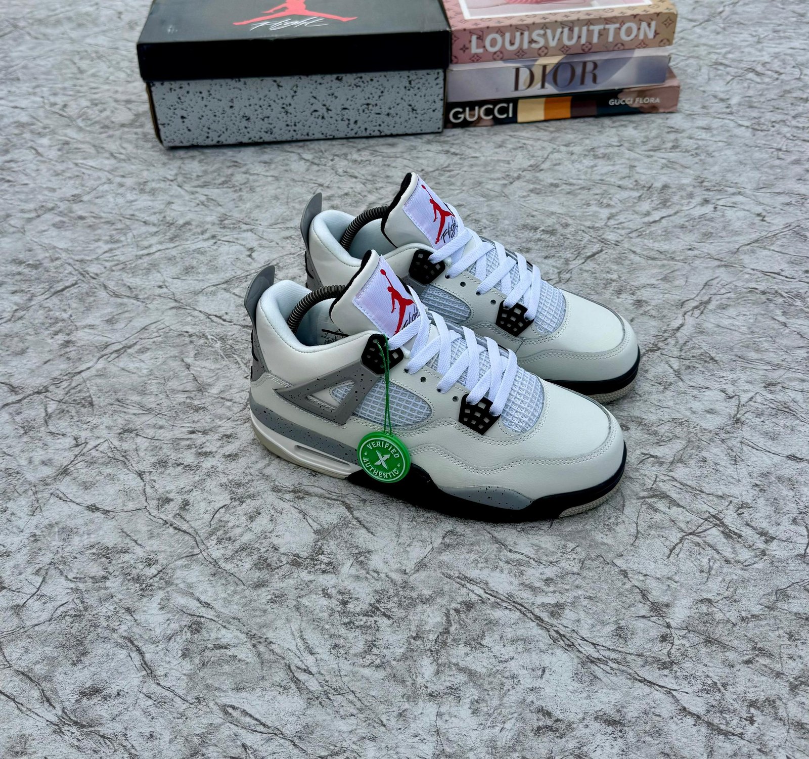 A pair of white and grey Air Jordan 4 sneakers with black accents, displayed on a marble surface with a shoe box and designer book covers in the background.