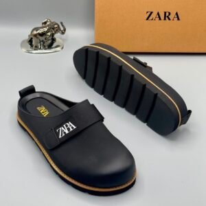 A pair of black leather clogs with yellow stitching and a ZARA logo, displayed on a white surface with a brown box and silver elephant figurine in the background.