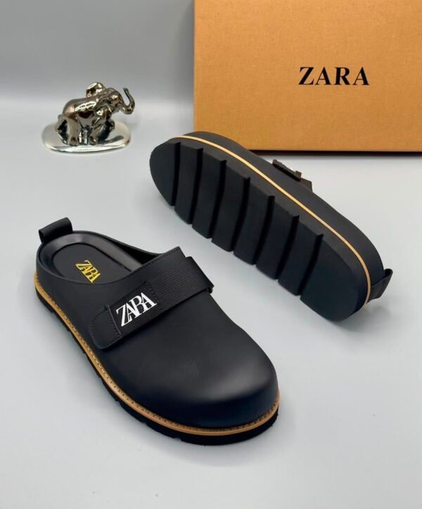 A pair of black leather clogs with yellow stitching and a ZARA logo, displayed on a white surface with a brown box and silver elephant figurine in the background.