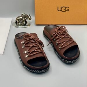A pair of black leather UGG sandals with silver laces and stitching, displayed with a UGG box and a decorative elephant figurine.