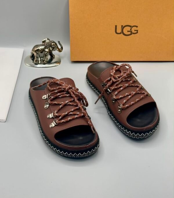 A pair of black leather UGG sandals with silver laces and stitching, displayed with a UGG box and a decorative elephant figurine.