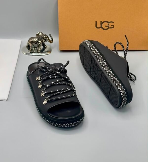 A pair of black leather UGG sandals with silver laces and stitching, displayed with a UGG box and a decorative elephant figurine.