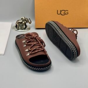 A pair of black leather UGG sandals with silver laces and stitching, displayed with a UGG box and a decorative elephant figurine.