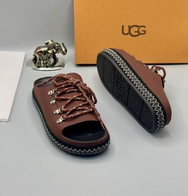 A pair of black leather UGG sandals with silver laces and stitching, displayed with a UGG box and a decorative elephant figurine.