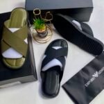 A pair of olive green and black cross strap sliders on a white surface, with a black shoe box, a small potted plant, and a black cloth bag with Emporio Armani logo.