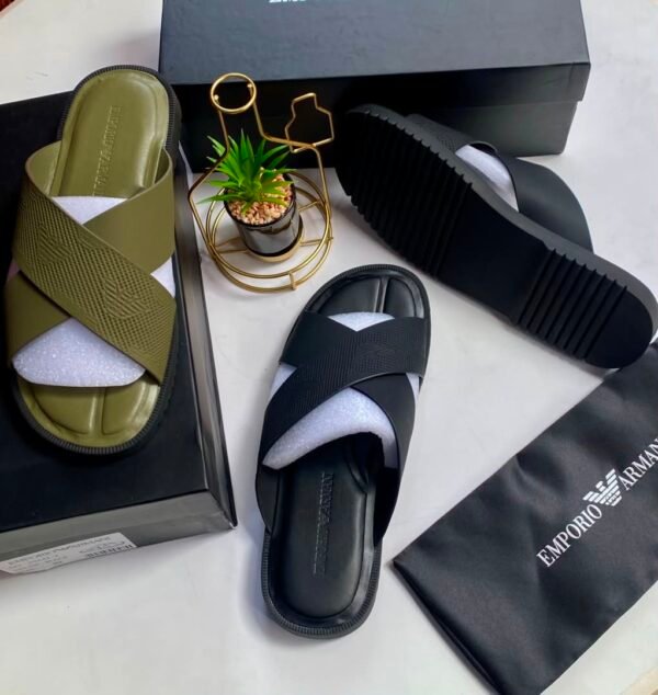 A pair of olive green and black cross strap sliders on a white surface, with a black shoe box, a small potted plant, and a black cloth bag with Emporio Armani logo.