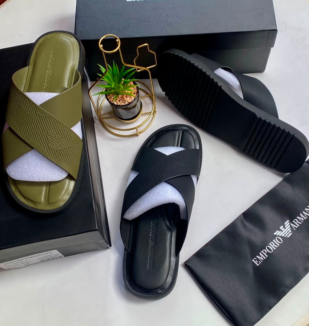 A pair of olive green and black cross strap sliders on a white surface, with a black shoe box, a small potted plant, and a black cloth bag with Emporio Armani logo.