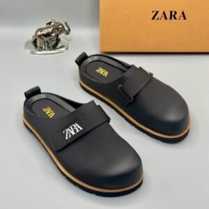 A pair of black leather clogs with yellow stitching and a ZARA logo, displayed on a white surface with a brown box and silver elephant figurine in the background.