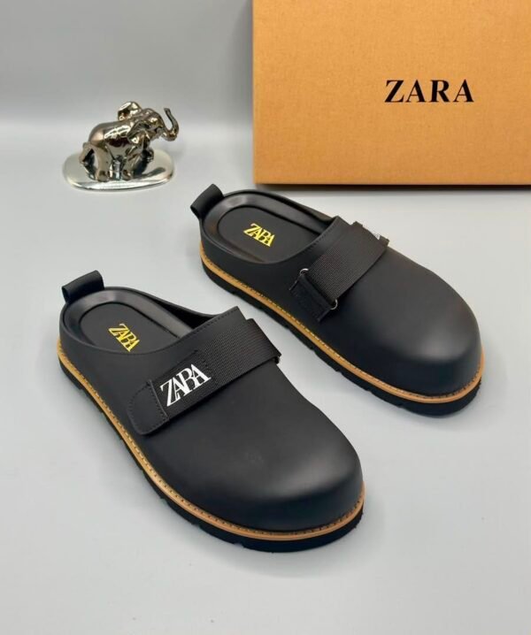 A pair of black leather clogs with yellow stitching and a ZARA logo, displayed on a white surface with a brown box and silver elephant figurine in the background.