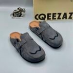 A pair of grey suede double-buckle moccasin shoes on a white background, with a cardboard box and metal elephant figurine nearby.