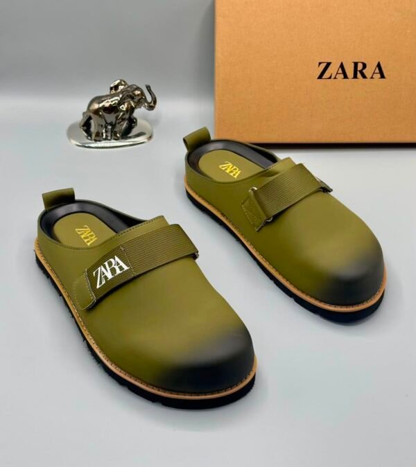 root A pair of olive green strap shoes with a silver elephant figurine and Zara box in the background.