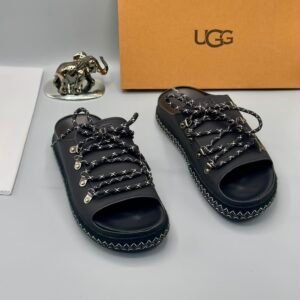 A pair of black leather UGG sandals with silver laces and stitching, displayed with a UGG box and a decorative elephant figurine.