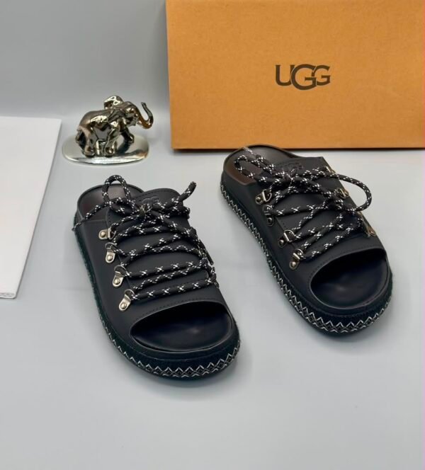 A pair of black leather UGG sandals with silver laces and stitching, displayed with a UGG box and a decorative elephant figurine.