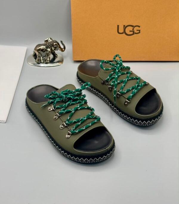 A pair of black leather UGG sandals with silver laces and stitching, displayed with a UGG box and a decorative elephant figurine.
