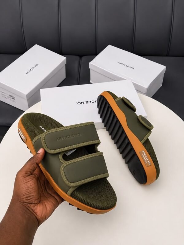 A pair of grey and orange sandals with two velcro straps, displayed on a white table with three white shoe boxes in the background.