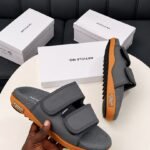 A pair of grey and orange sandals with two velcro straps, displayed on a white table with three white shoe boxes in the background.