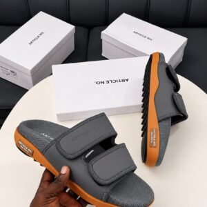 A pair of grey and orange sandals with two velcro straps, displayed on a white table with three white shoe boxes in the background.