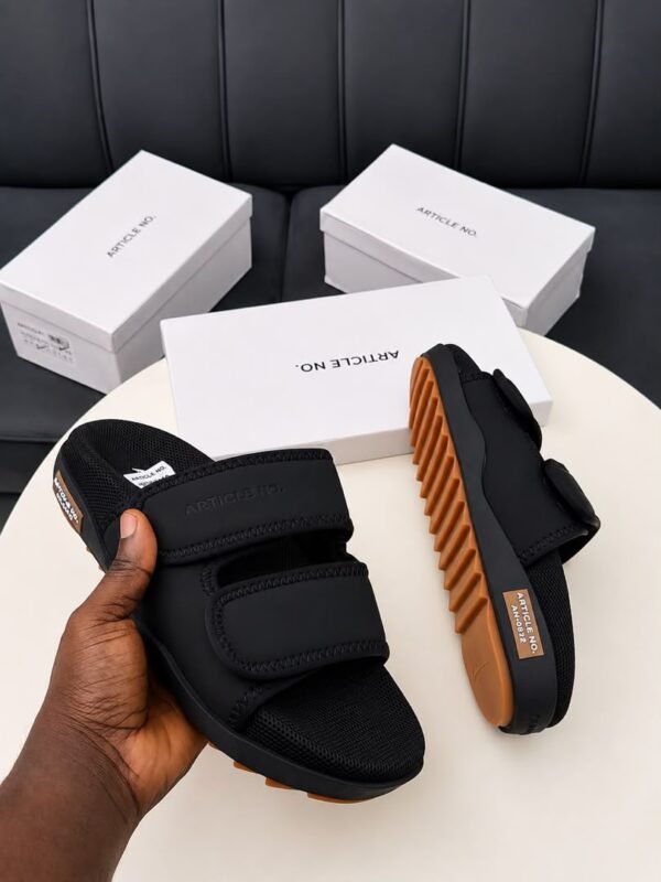 A pair of grey and orange sandals with two velcro straps, displayed on a white table with three white shoe boxes in the background.