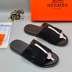 A pair of luxurious black sandals with rhinestone straps, displayed on a grey surface with an orange Hermes Paris box and a silver elephant figurine in the background.