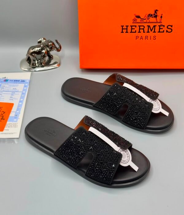 A pair of luxurious black sandals with rhinestone straps, displayed on a grey surface with an orange Hermes Paris box and a silver elephant figurine in the background.