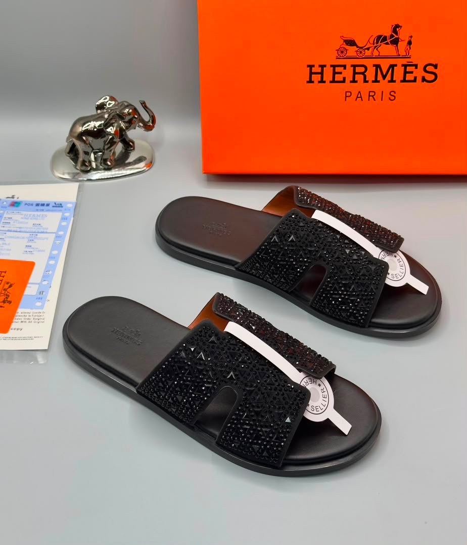 A pair of luxurious black sandals with rhinestone straps, displayed on a grey surface with an orange Hermes Paris box and a silver elephant figurine in the background.