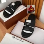 A pair of black slides with white inserts and a logo on the strap, placed in a brown shoe box with a white interior and a Loro Piana logo on the lid, alongside a small red toy motorcycle.