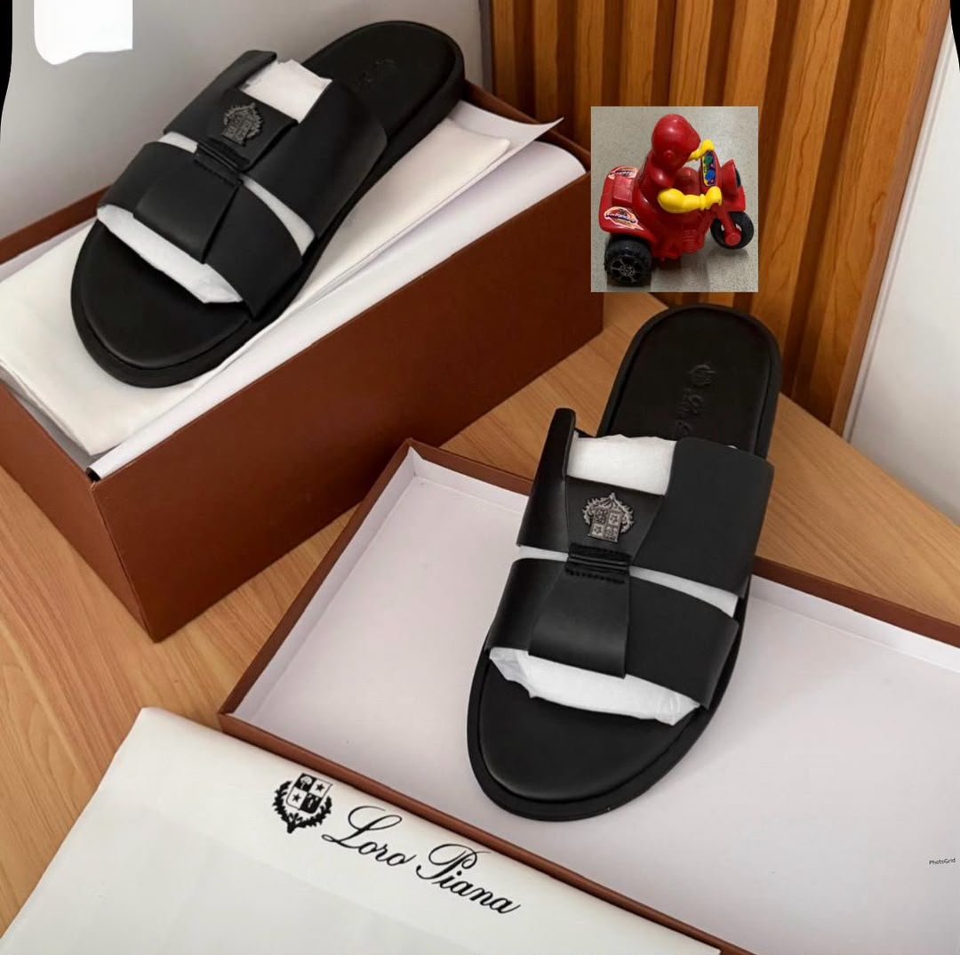 A pair of black slides with white inserts and a logo on the strap, placed in a brown shoe box with a white interior and a Loro Piana logo on the lid, alongside a small red toy motorcycle.